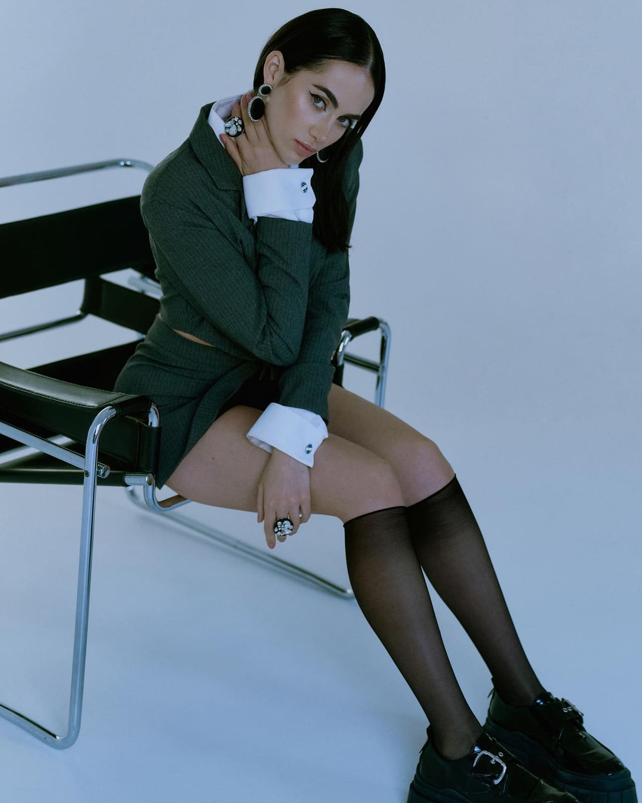 Abel in a green blazer and skirt, sitting on a black leather chair.