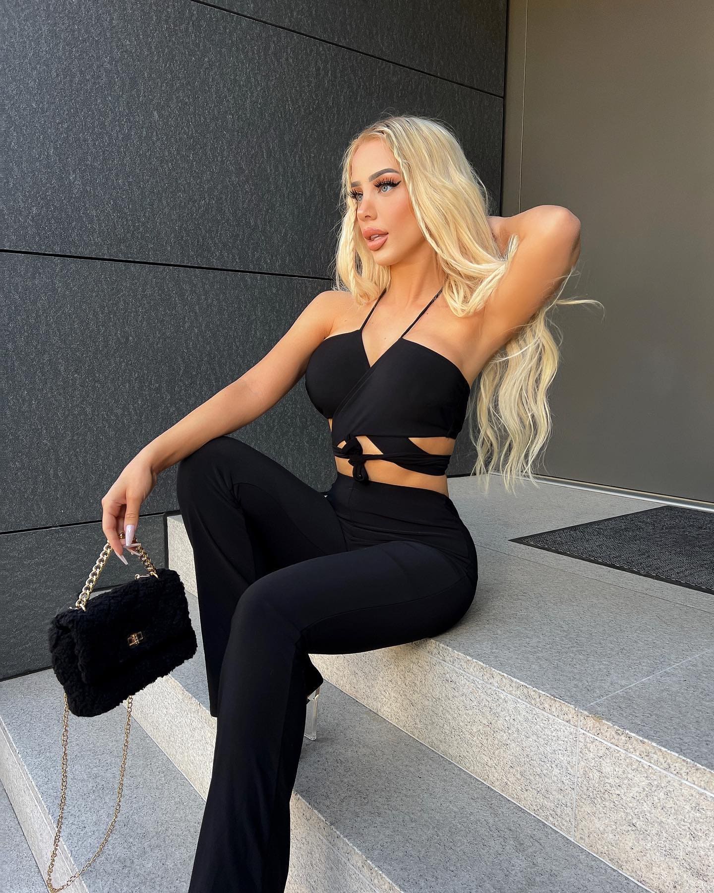 A blonde woman sits on a stone step before a dark wall, wearing a black strappy crop top and flared trousers, with one arm raised behind her head.