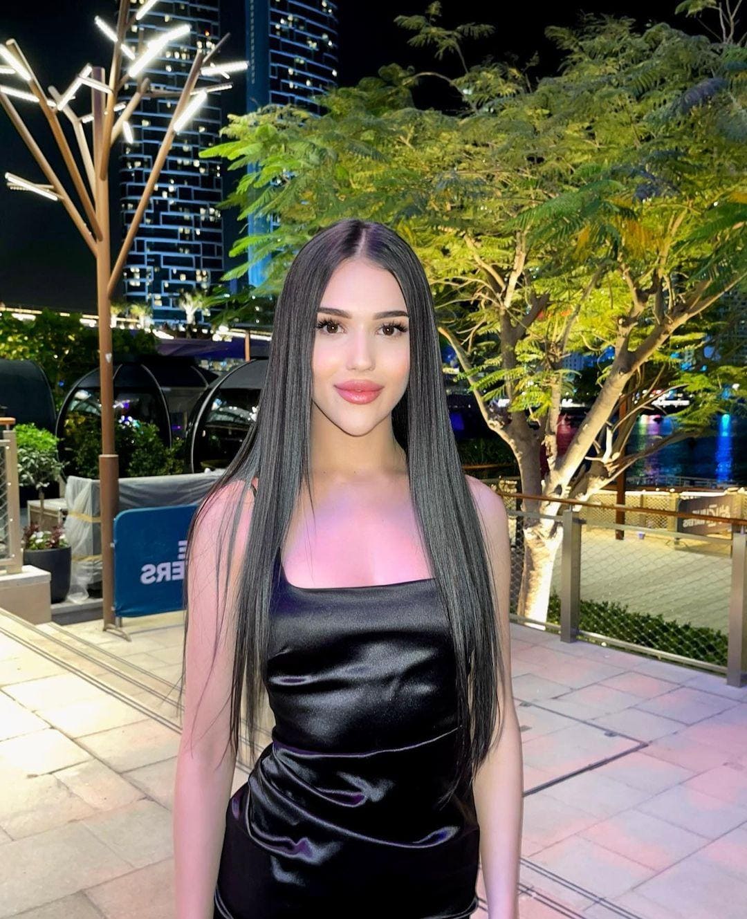 Odessa stands on a paved walkway at night in a black satin sleeveless top, with illuminated trees and a high-rise building behind her.