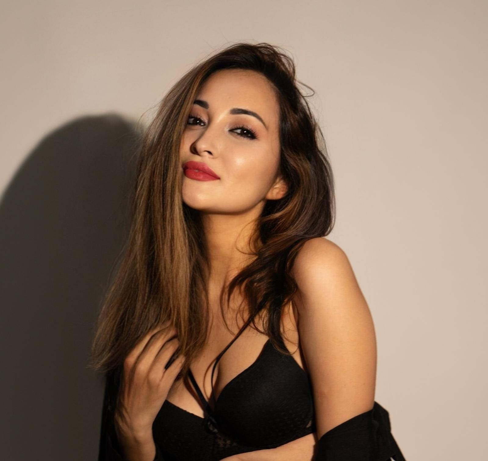 Zara with long brown hair wears a black bra and draped garment, posing with a hand near her chest against a plain light background.