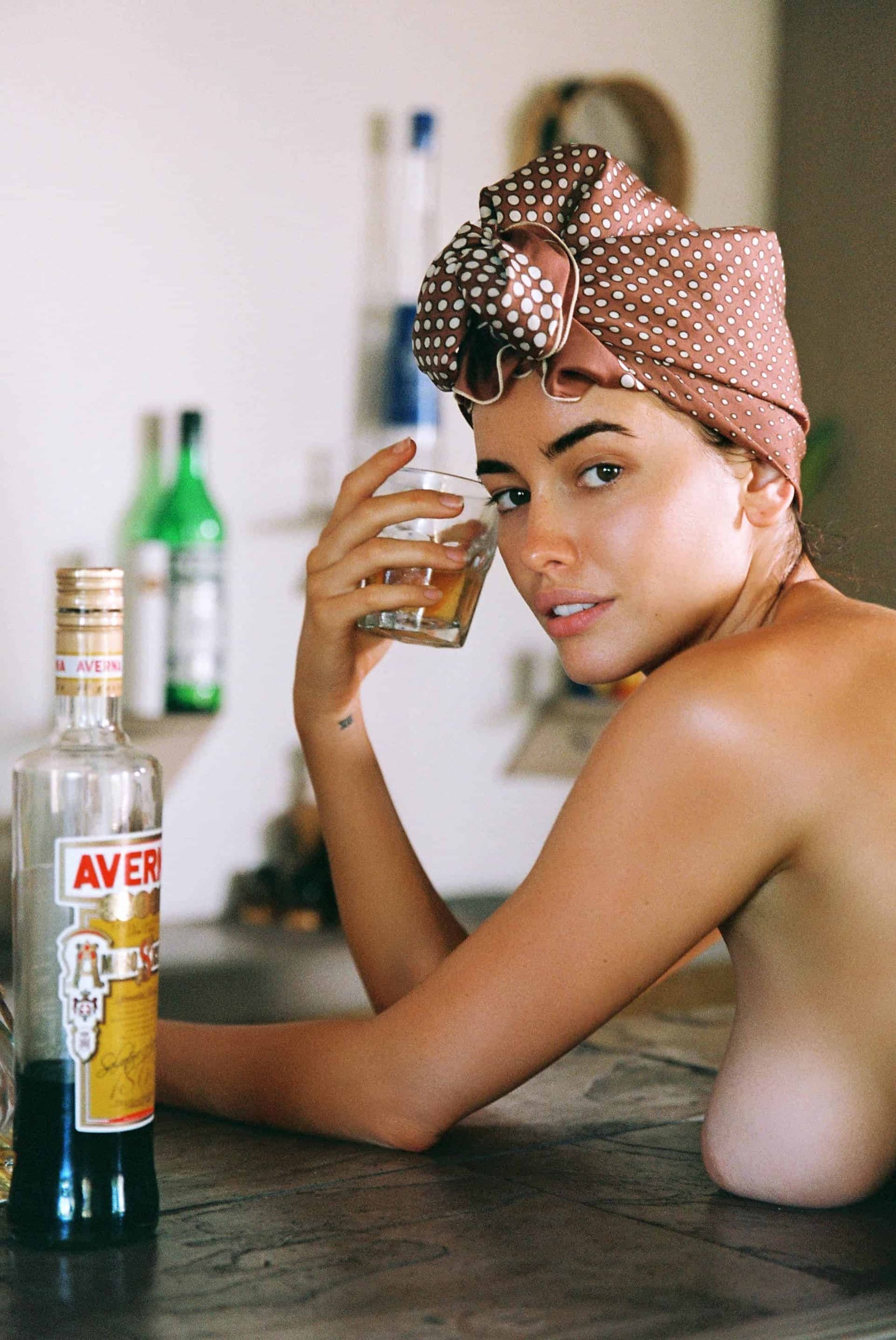 A woman in a polka-dot headscarf leans on a table holding a glass of amber liquid.