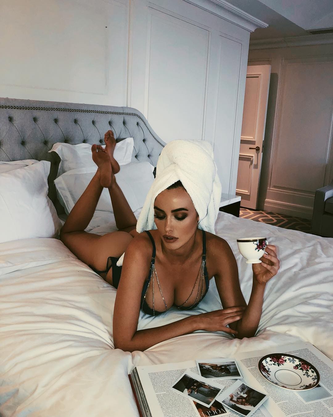 A woman in a black strappy bra lies on a bed holding a teacup, with a magazine nearby.