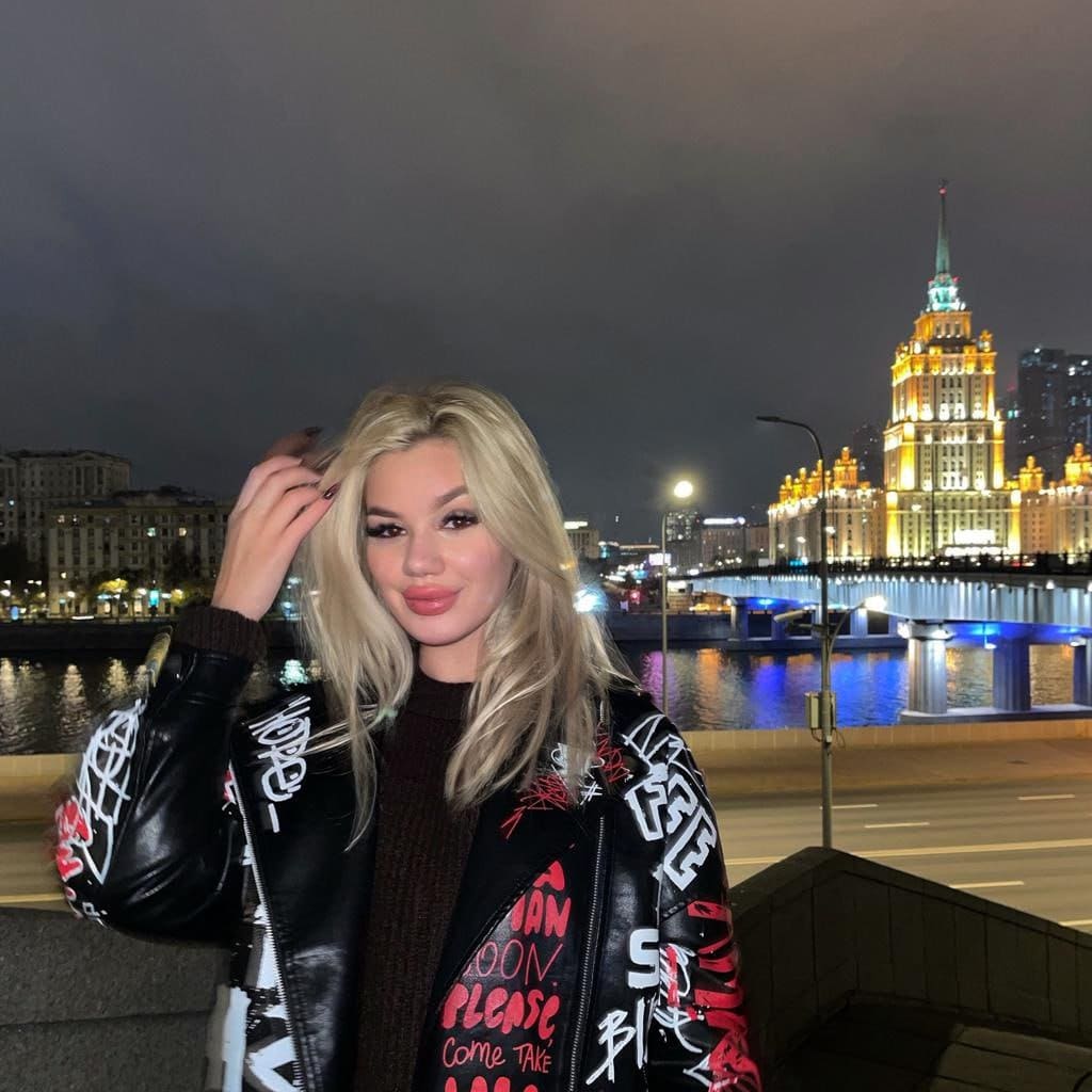 Sonya stands outdoors at night by a lit city skyline, wearing a black leather jacket with graffiti lettering and touching her long blonde hair.