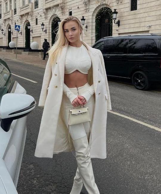 Kara stands on a city street in a long white coat over white pants and top, holding a white handbag near parked vehicles.