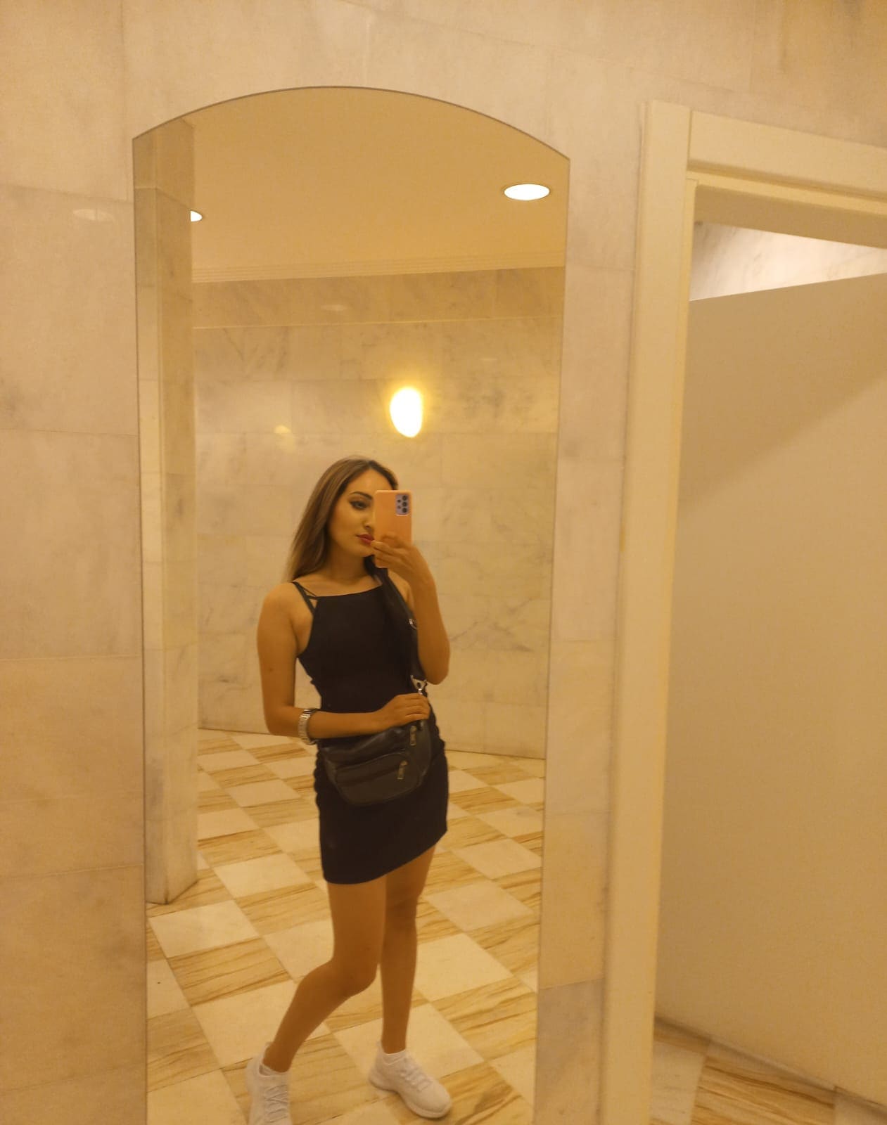 A mirror selfie shows a woman in a black mini dress and white sneakers holding a phone with an orange case in a marble-tiled room.