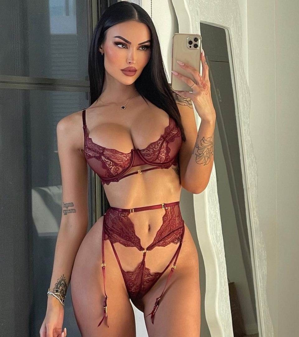 Mirror selfie of a woman in red lace lingerie with a garter belt, standing indoors near a window and white curtain.