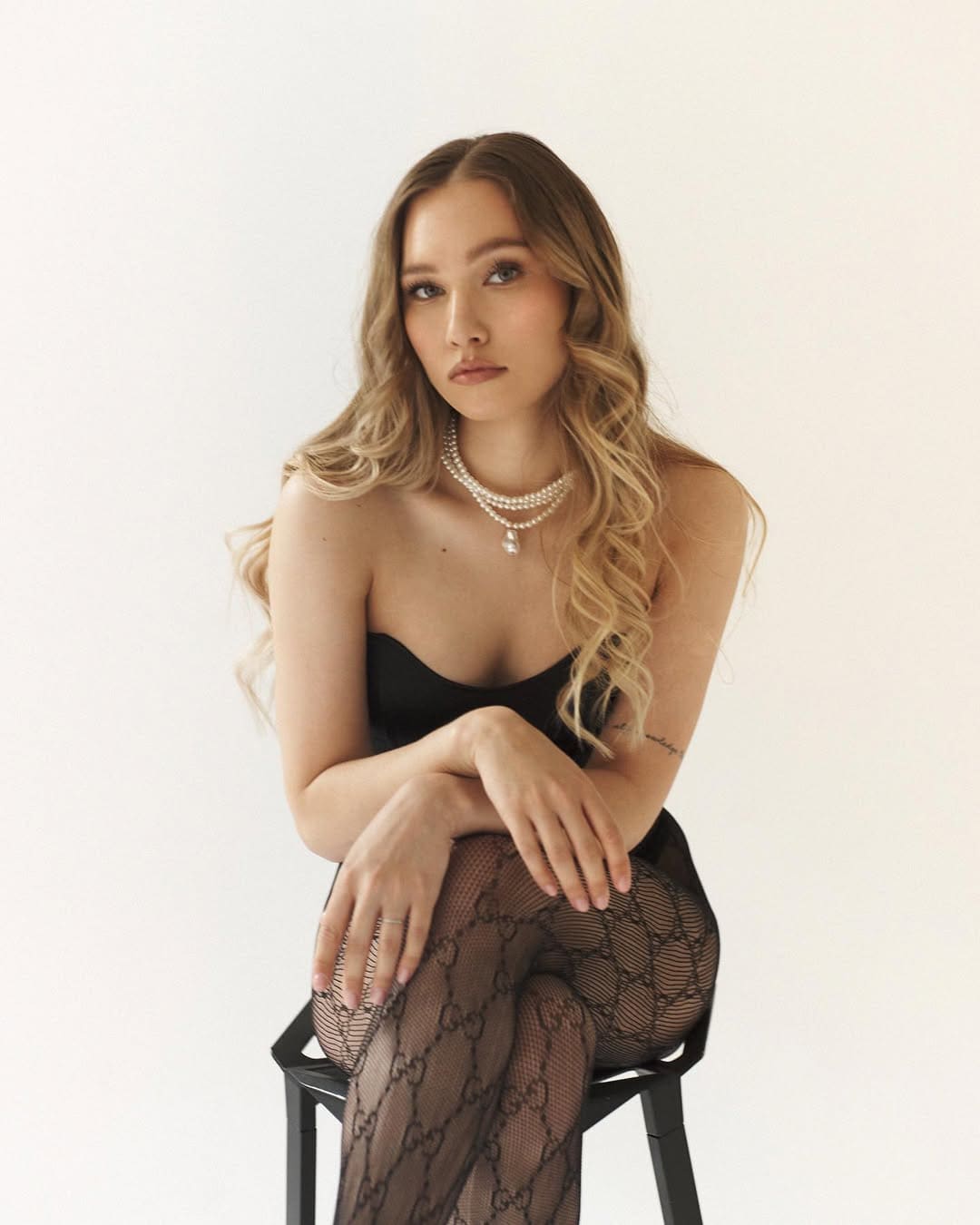 A woman with wavy blonde hair sits on a black stool in a black strapless top and sheer tights with a pearl necklace.