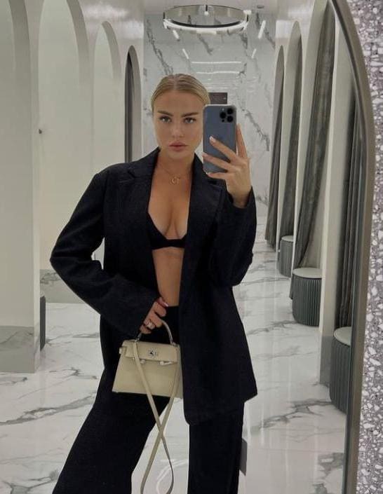 Mirror selfie in a marble hallway showing a woman in an open black blazer, black bra, and trousers holding a light bag.