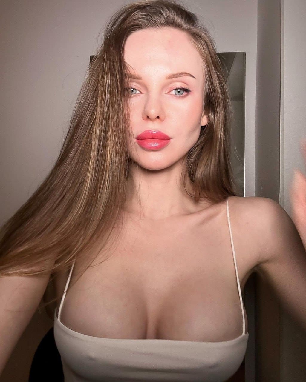 Selfie of a woman with light-brown hair in a thin-strapped light top, standing in a room with a plain wall and a mirror visible behind her.
