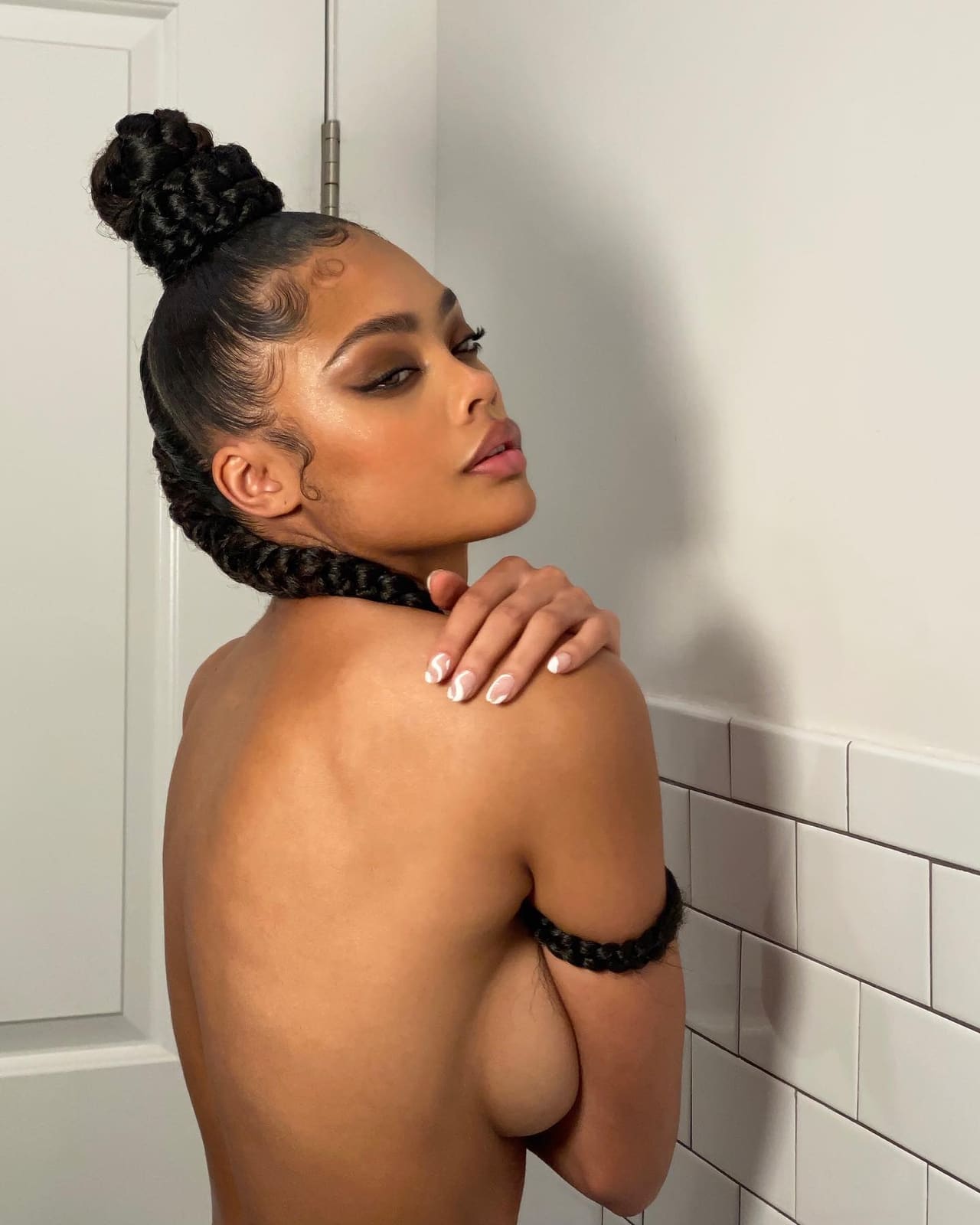 A woman with hair in a high bun stands bare-backed against a white tiled wall, her hand resting on her shoulder.