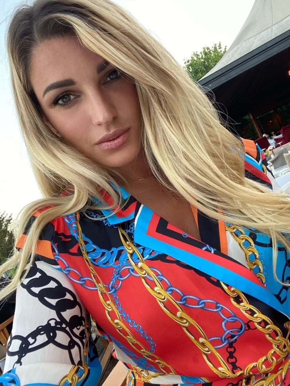 A woman with long blonde hair in a patterned blouse poses outdoors near a tented structure.
