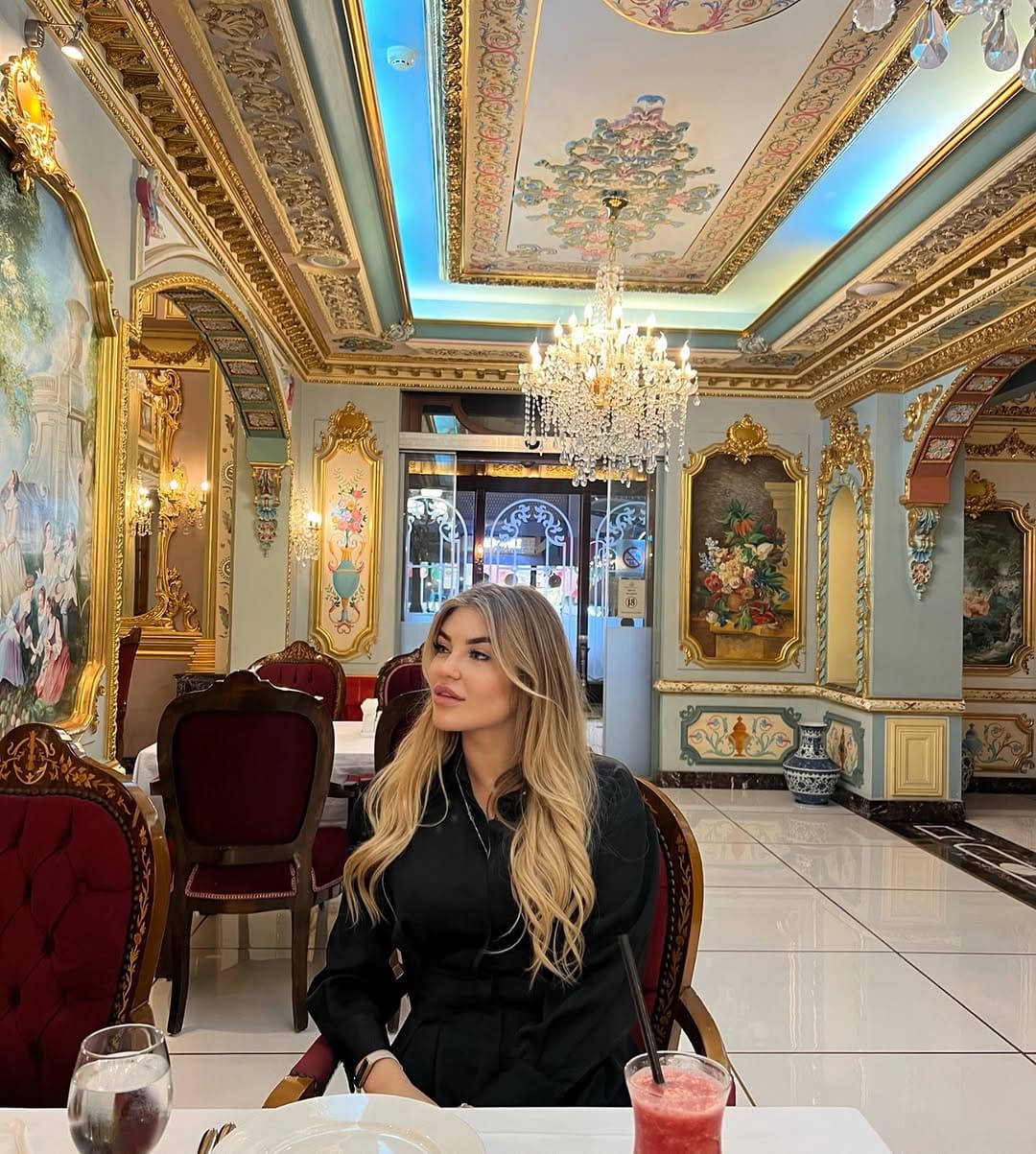 A woman in a black long-sleeved top sits at a white-clothed table in an ornate restaurant, looking to the side under a crystal chandelier.