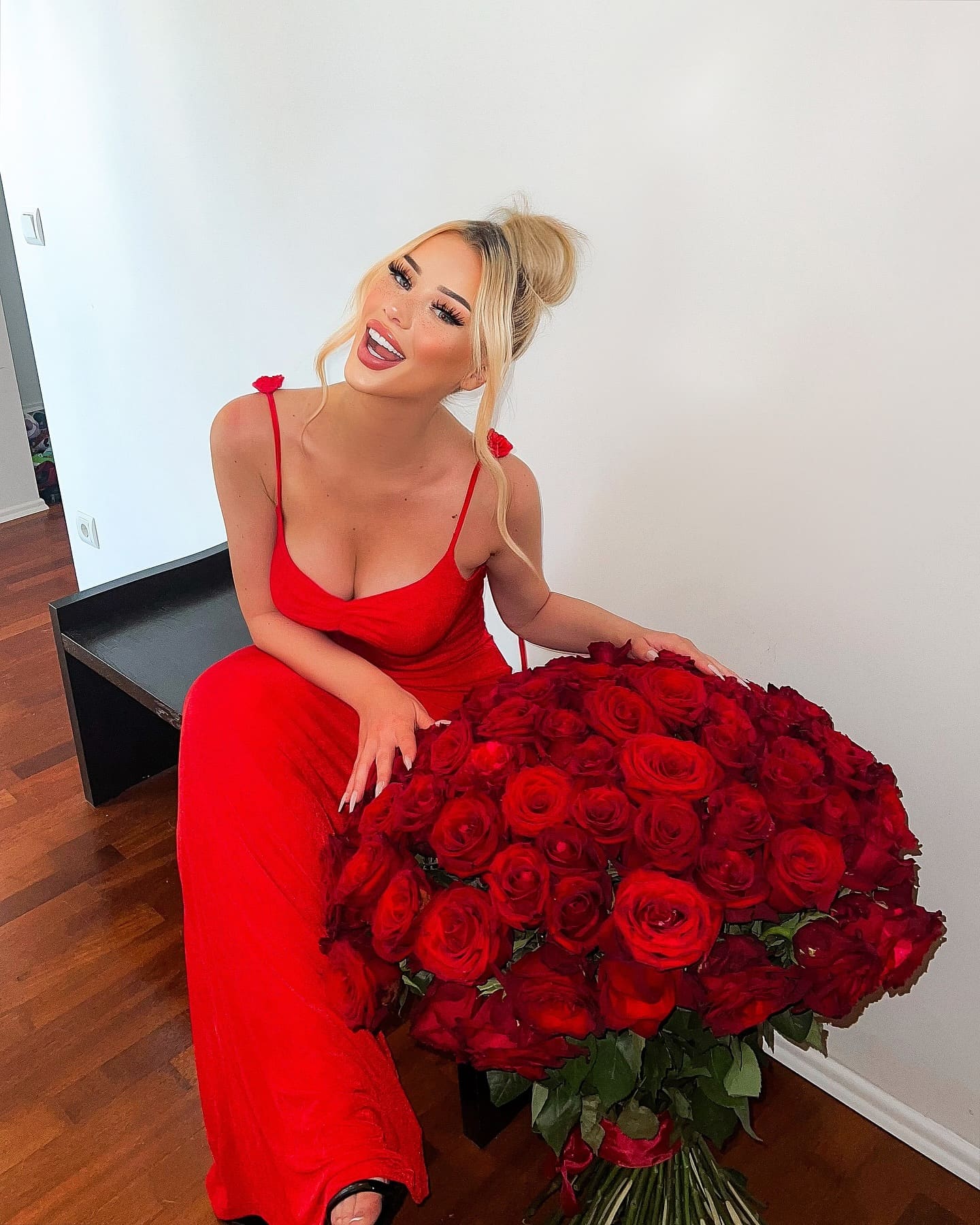 A blonde woman sits on a black bench against a white wall, wearing a red spaghetti-strap dress and holding a large bouquet of red roses.