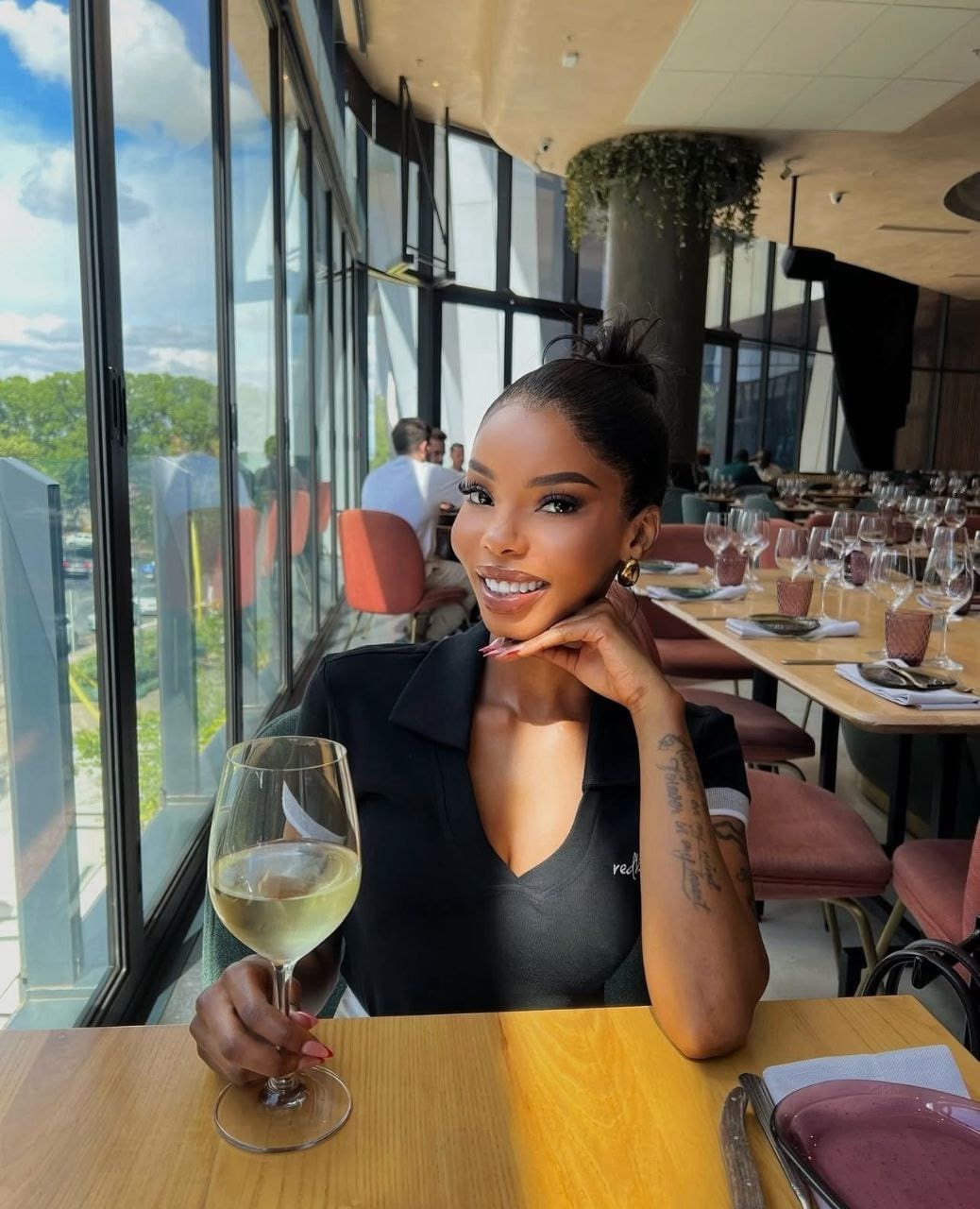 Liliana sits at a restaurant table in a black V-neck top, holding white wine with her chin resting on her hand, city view through windows behind.