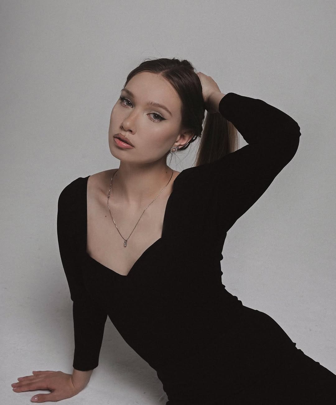 Alexandra in a black V-neck dress with a ponytail, posing with one hand behind her head against a light background.