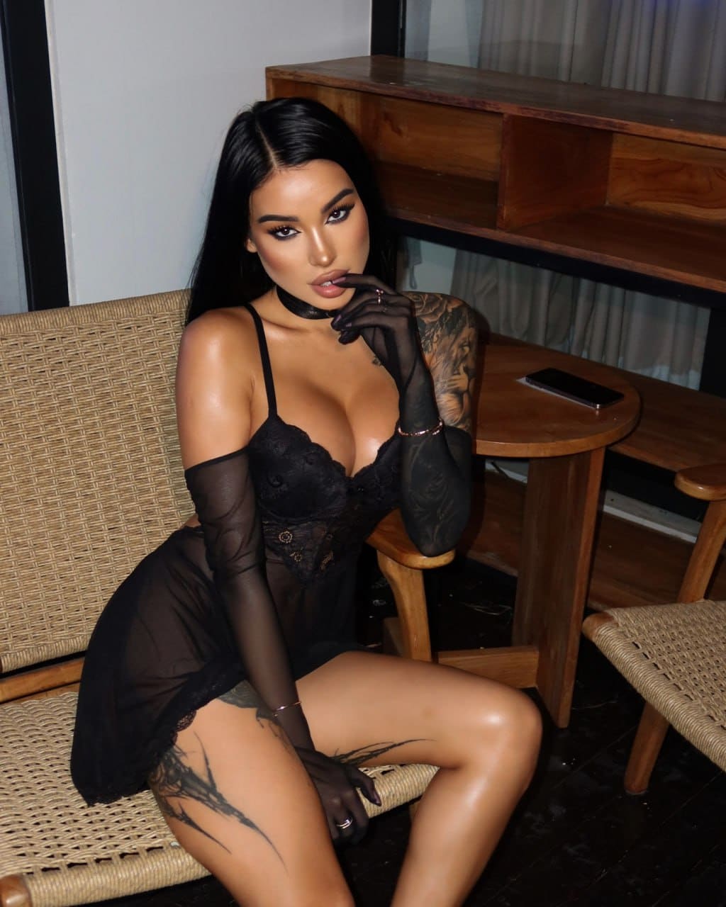A woman with long dark hair sits on a woven chair in a sheer black lace bodysuit and long gloves, hand raised to her chin.